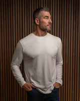 A man wearing a fitted, long-sleeve crewneck t-shirt in a light gray color, paired with dark blue jeans, standing against a dark brown wood slat wall.