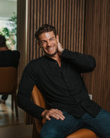 A smiling man in a black long-sleeve button-up shirt and blue jeans sits on a tan leather chair in front of a dark wood slat wall.