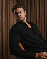 A man wearing a black long-sleeve button-up shirt sits in a chair against a dark wood slat wall background.