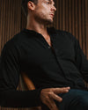 A man wearing a black long-sleeve button-up shirt and dark pants, seated in a leather chair against a warm wood slat wall.