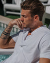 A man wearing a white short-sleeve polo shirt, leaning on a poolside lounge chair with a blurred outdoor background.