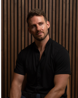 A man wearing a black long-sleeve button-up shirt and dark blue jeans, standing in front of a rustic wooden plank wall.