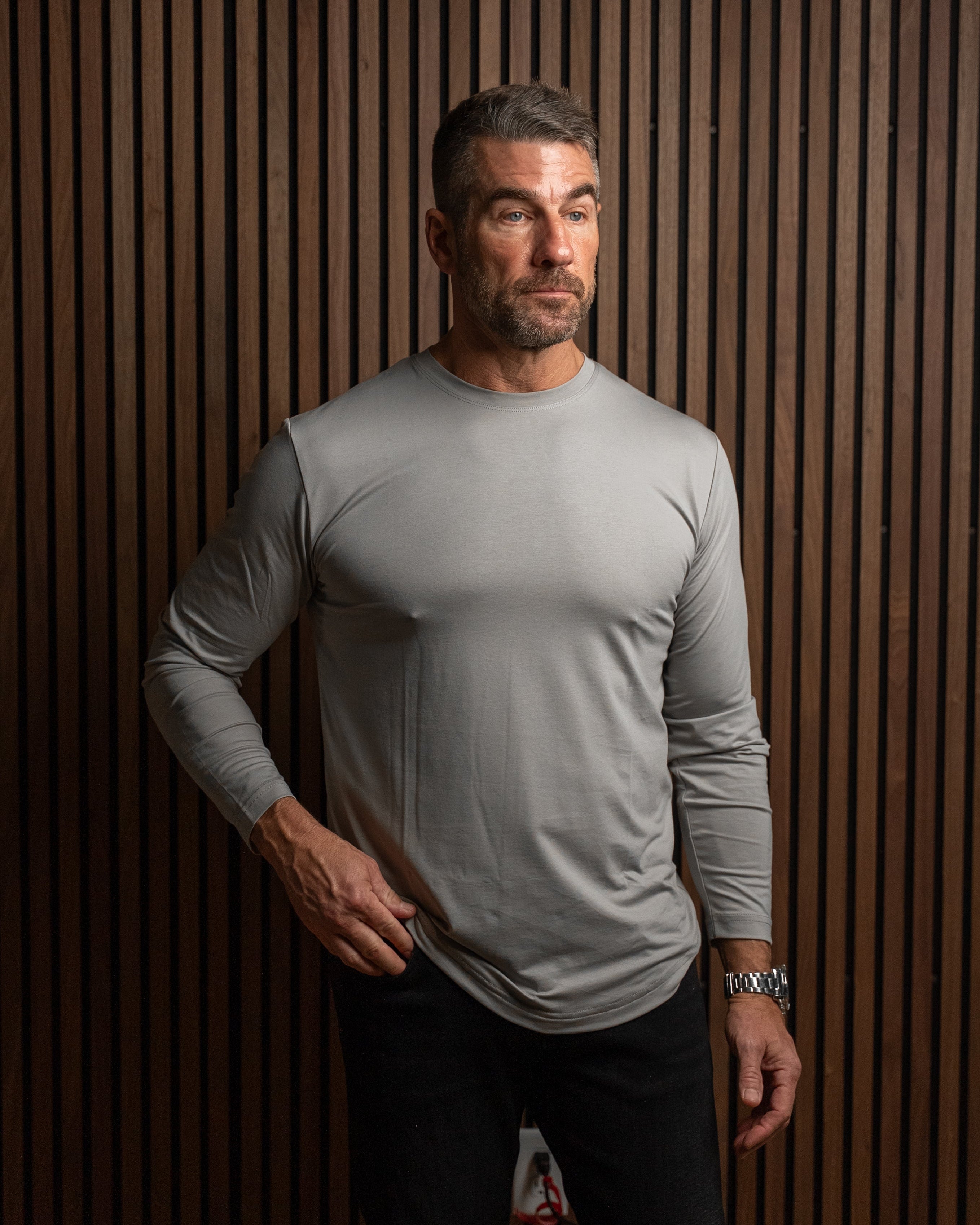 A man wearing a light gray long-sleeve athletic shirt and black pants, standing against a dark brown vertically slatted wood wall.