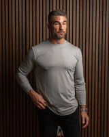 A man wearing a light gray long-sleeve athletic shirt and black pants, standing against a dark brown vertically slatted wood wall.