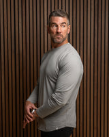 A man wearing a light gray long-sleeve athletic shirt and black pants, standing against a dark brown vertically slatted wooden wall.