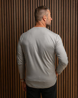 A man viewed from behind wearing a light gray long-sleeve crewneck shirt, standing against a dark brown vertical slat wall.