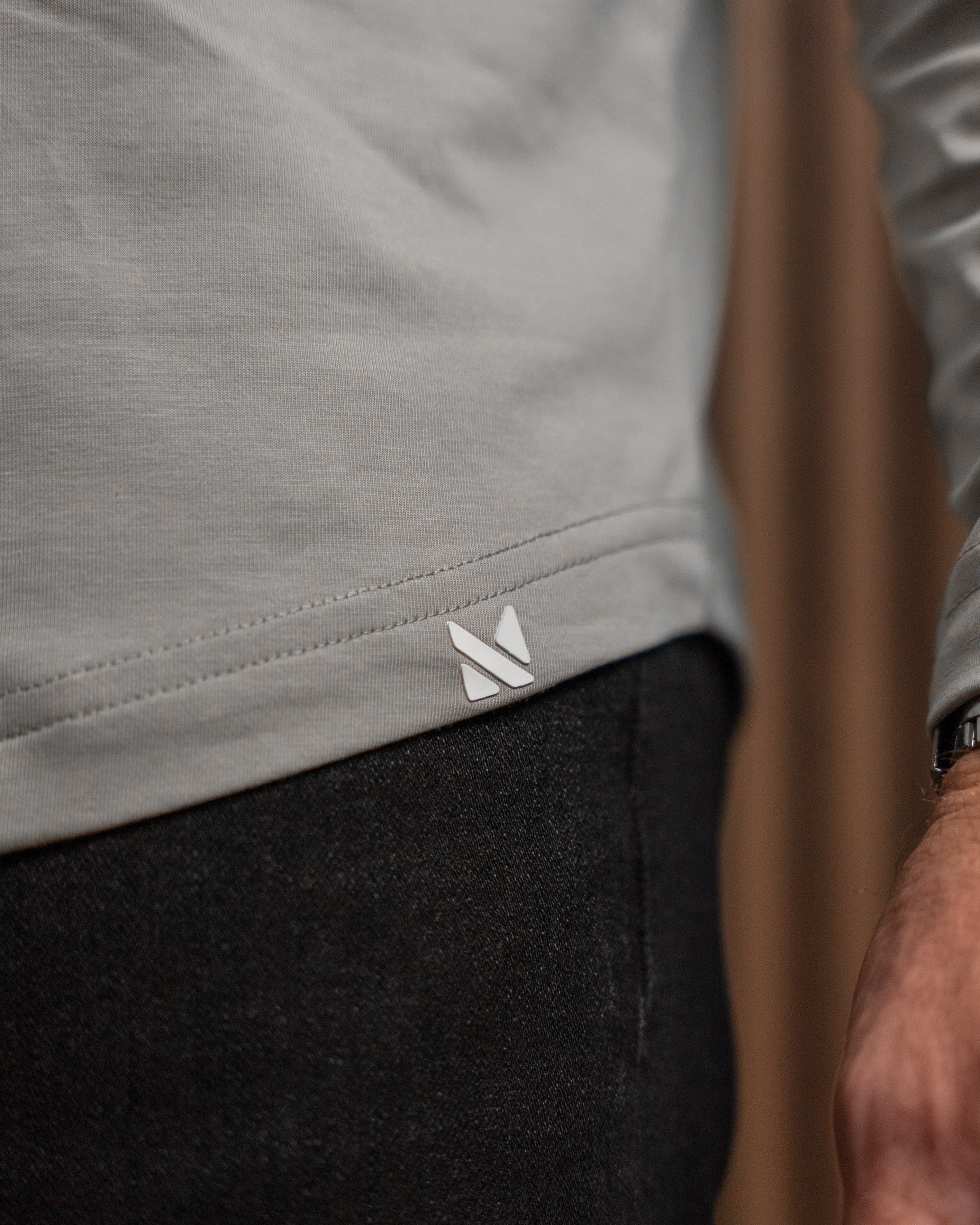 Close-up of a person wearing a light gray long-sleeve top and dark charcoal pants, with a small white geometric logo patch near the hem of the shirt.