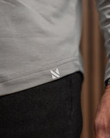 Close-up of a person wearing a light gray long-sleeve top and dark charcoal pants, with a small white geometric logo patch near the hem of the shirt.