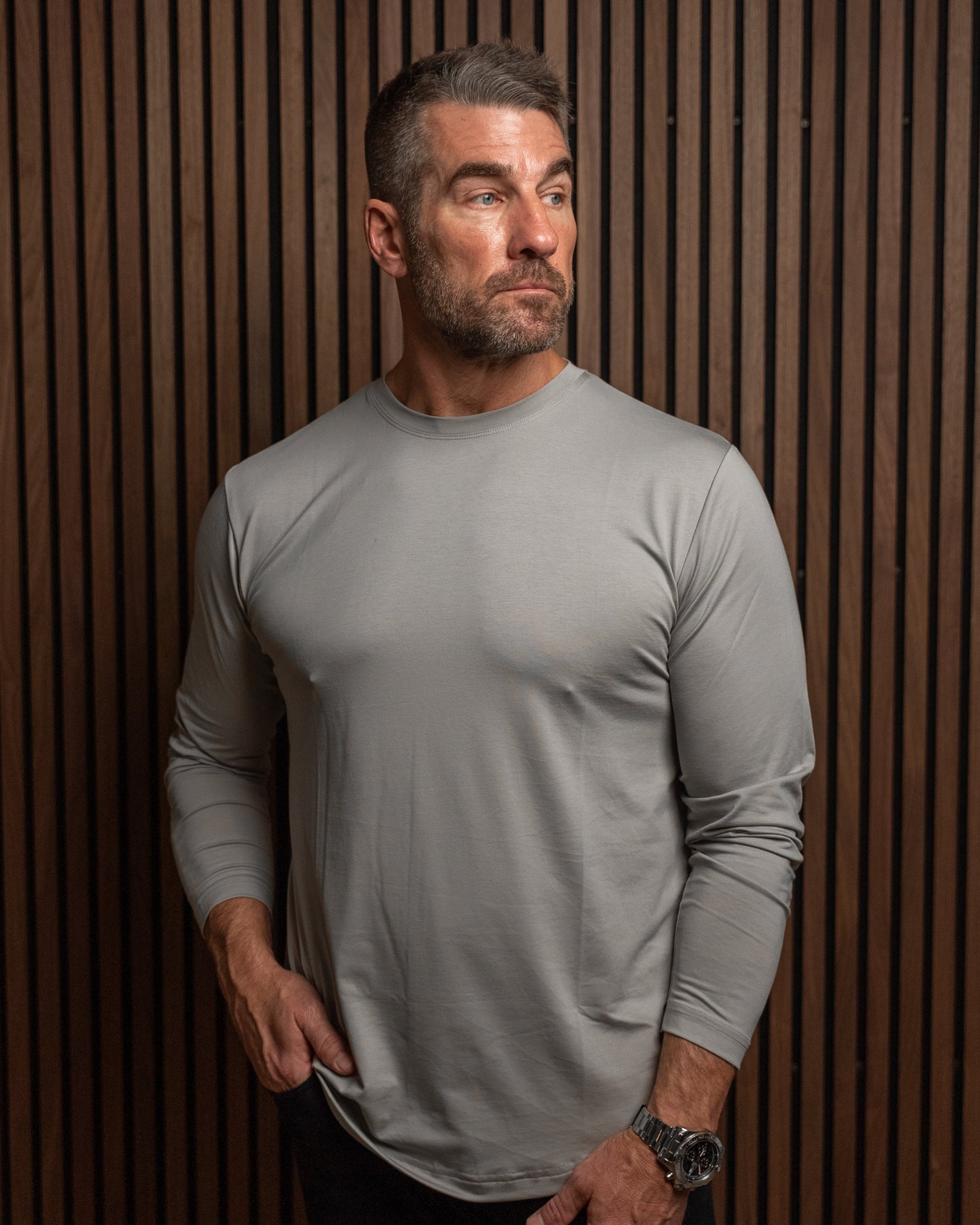 A muscular man wearing a tight-fitting, light gray long-sleeve athletic shirt and dark pants, standing against a dark brown wood slat wall.