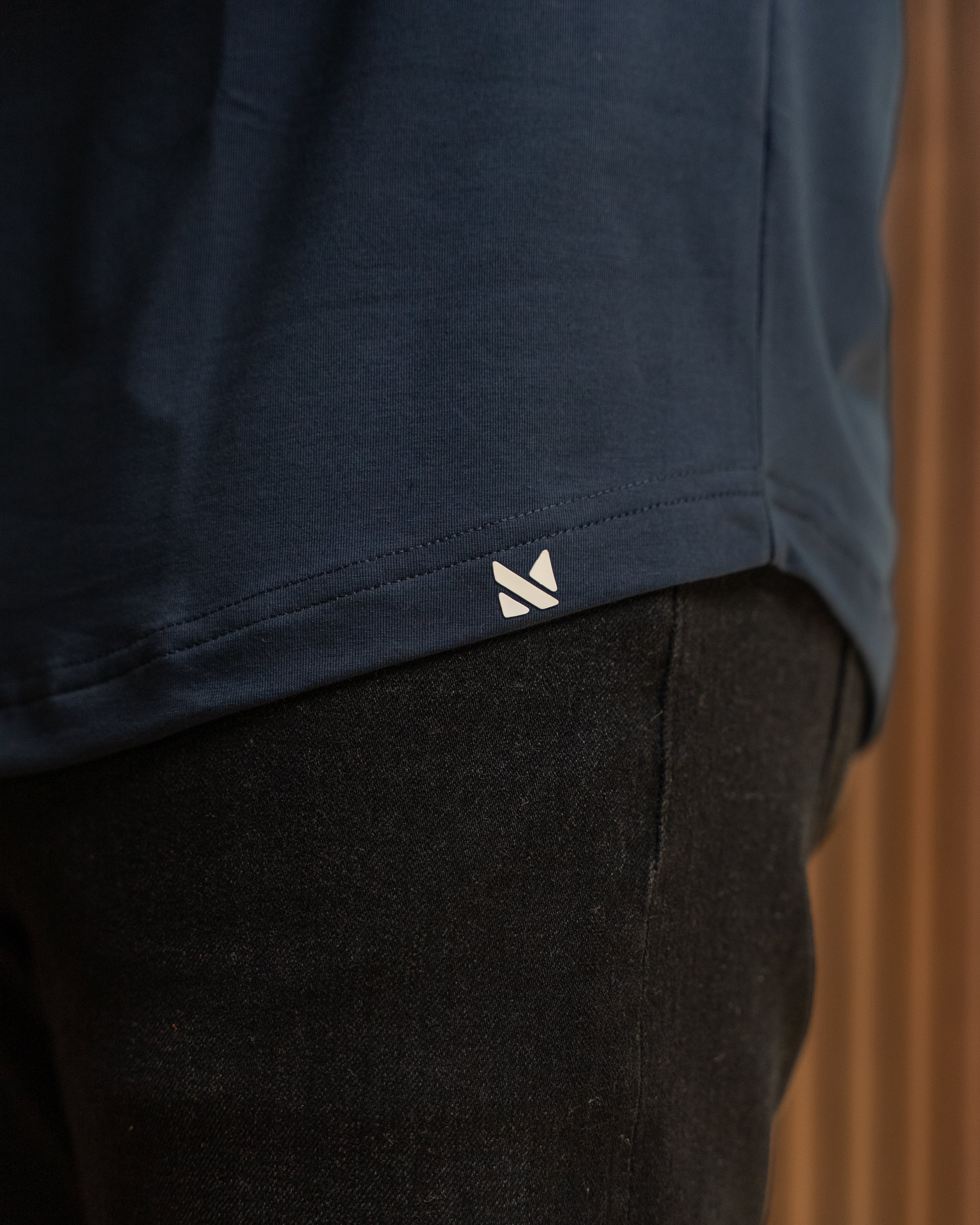 Close-up of a navy blue top with a small white geometric logo near the hem, worn with dark pants against a warm brown background.