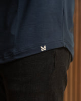 Close-up of a navy blue top with a small white geometric logo near the hem, worn with dark pants against a warm brown background.