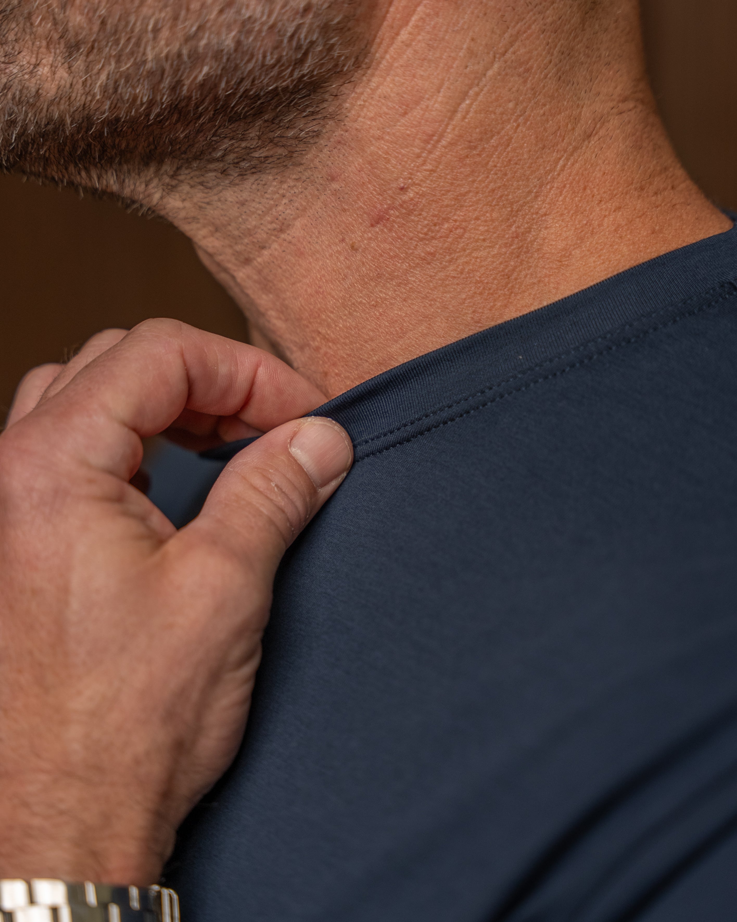 Close-up of a man's neck and shoulder wearing a dark navy blue t-shirt, with his hand pulling the collar slightly outward to show the fit and neckline construction.