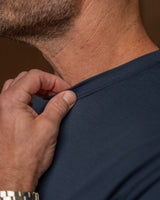 Close-up of a man's neck and shoulder wearing a dark navy blue t-shirt, with his hand pulling the collar slightly outward to show the fit and neckline construction.