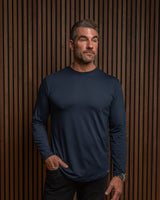 A man wearing a dark navy blue long-sleeve crewneck shirt, standing against a dark brown vertical wood slat wall.