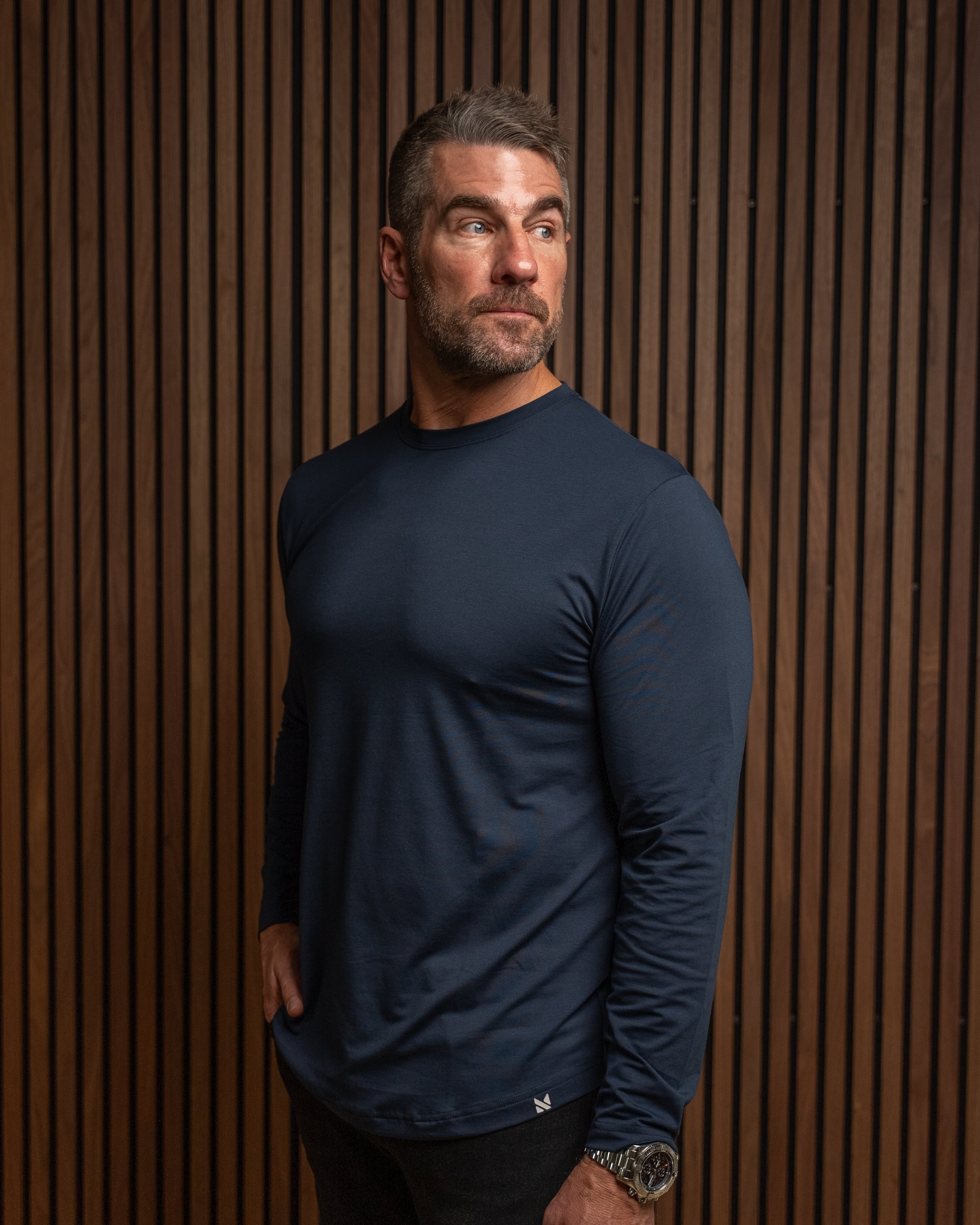 A man wearing a navy blue long-sleeve athletic shirt and black pants, standing against a brown wood slat wall.