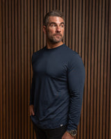 A man wearing a navy blue long-sleeve athletic shirt and black pants, standing against a brown wood slat wall.