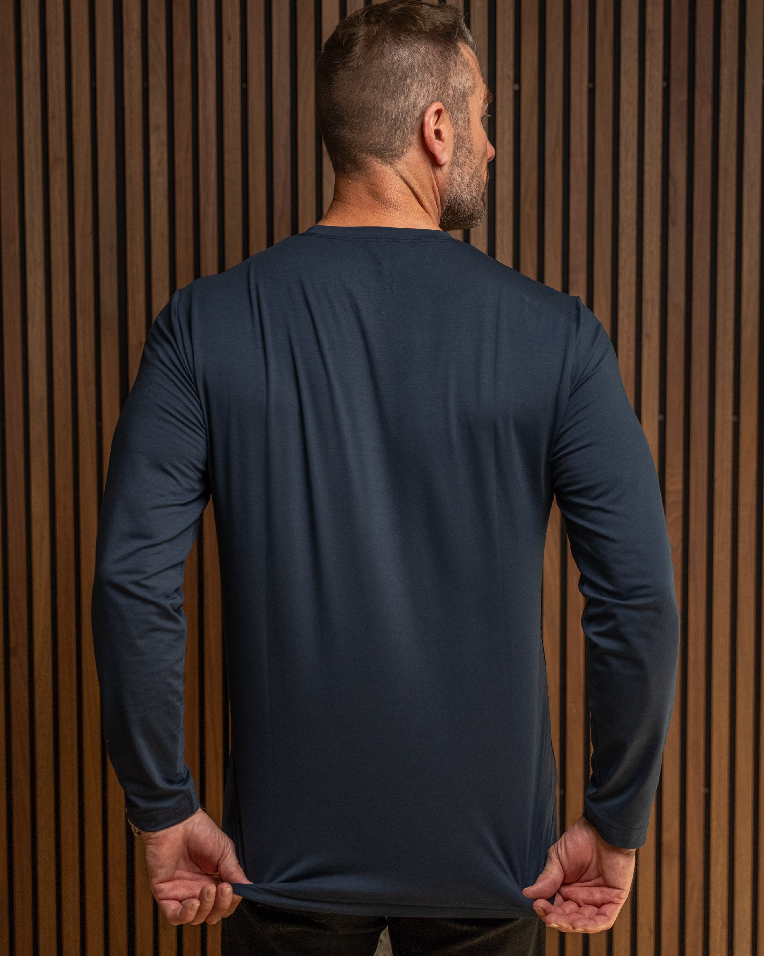 A man viewed from behind wearing a dark navy long-sleeve athletic shirt, stretching the fabric at the waist against a brown wood slat wall.