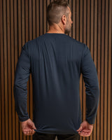 A man viewed from behind wearing a dark navy long-sleeve athletic shirt, stretching the fabric at the waist against a brown wood slat wall.