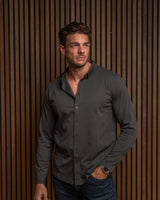 A man wearing a dark charcoal gray long-sleeve button-down shirt and blue jeans, standing against a brown vertical slat wall.
