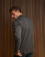 A man wearing a dark gray long-sleeve blazer and blue jeans, shown from a side-back angle against a vertical wood slat wall.