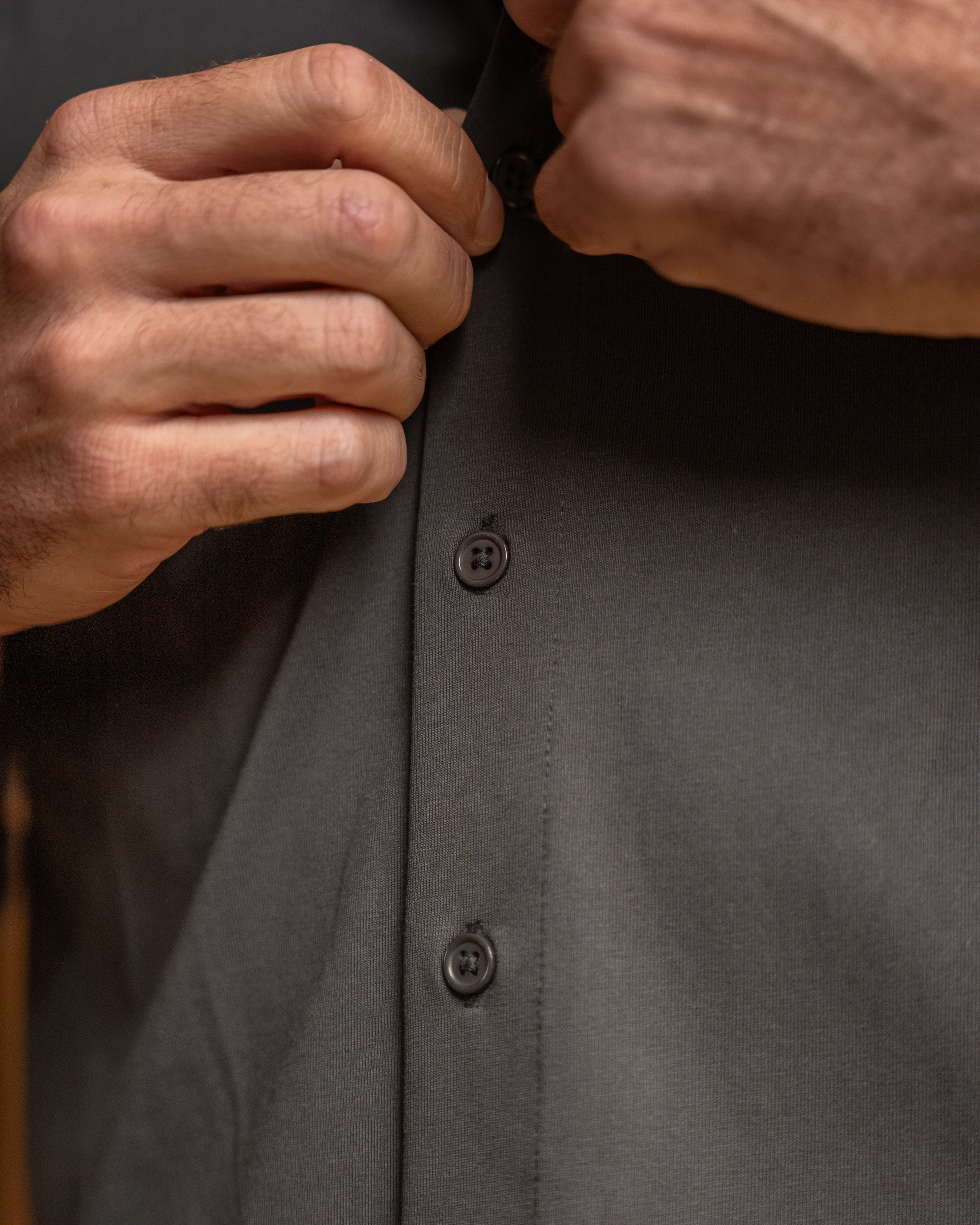 Close-up of a person's hands buttoning a dark charcoal gray long-sleeve dress shirt.