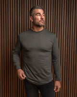 A man wearing a dark gray long-sleeve crewneck shirt and dark jeans, standing against a brown vertical slat wall.