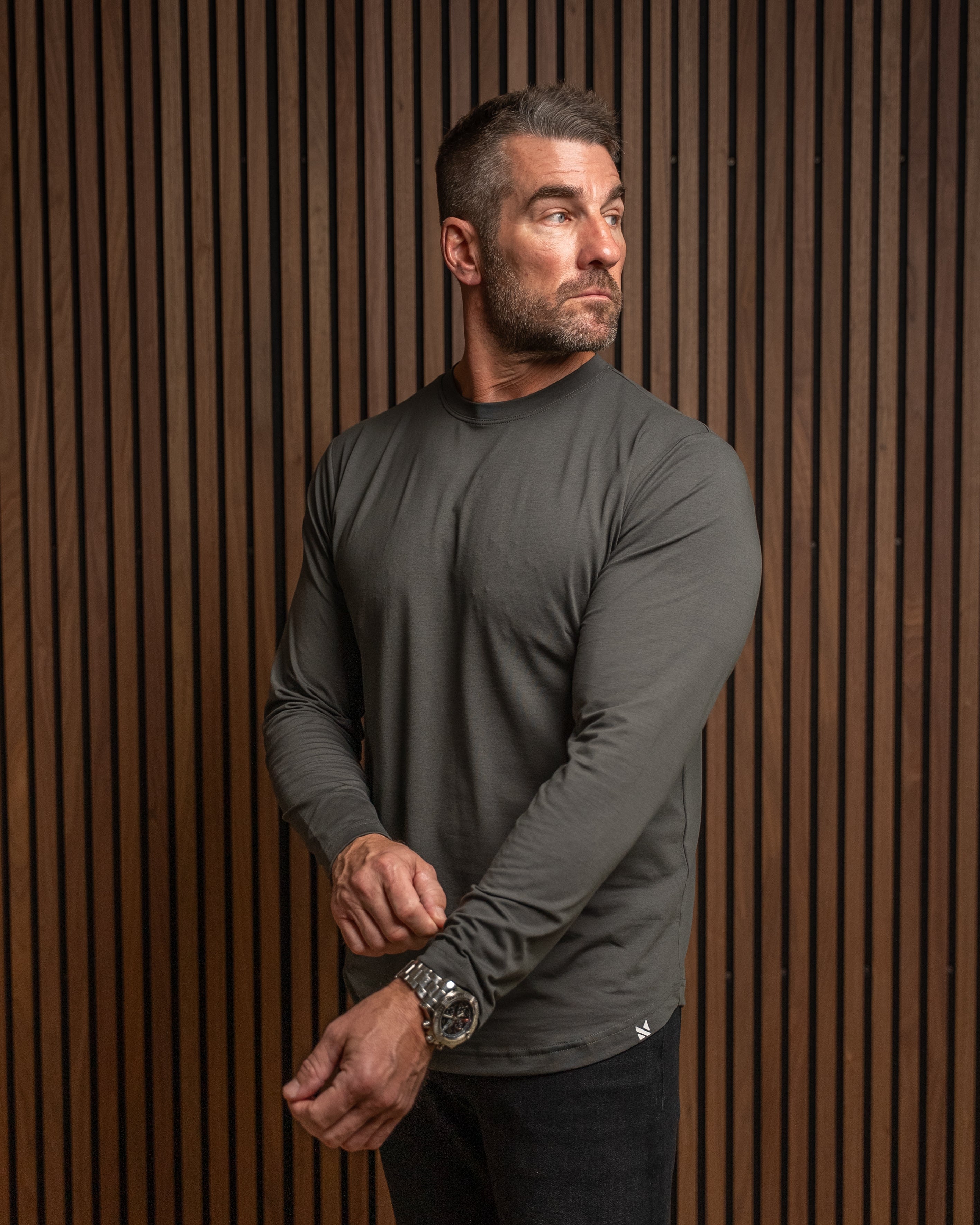 A man wearing a dark gray long-sleeve athletic shirt and black pants, standing against a brown vertical slat wall.