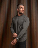 A man wearing a dark gray long-sleeve athletic shirt and black pants, standing against a brown vertical slat wall.