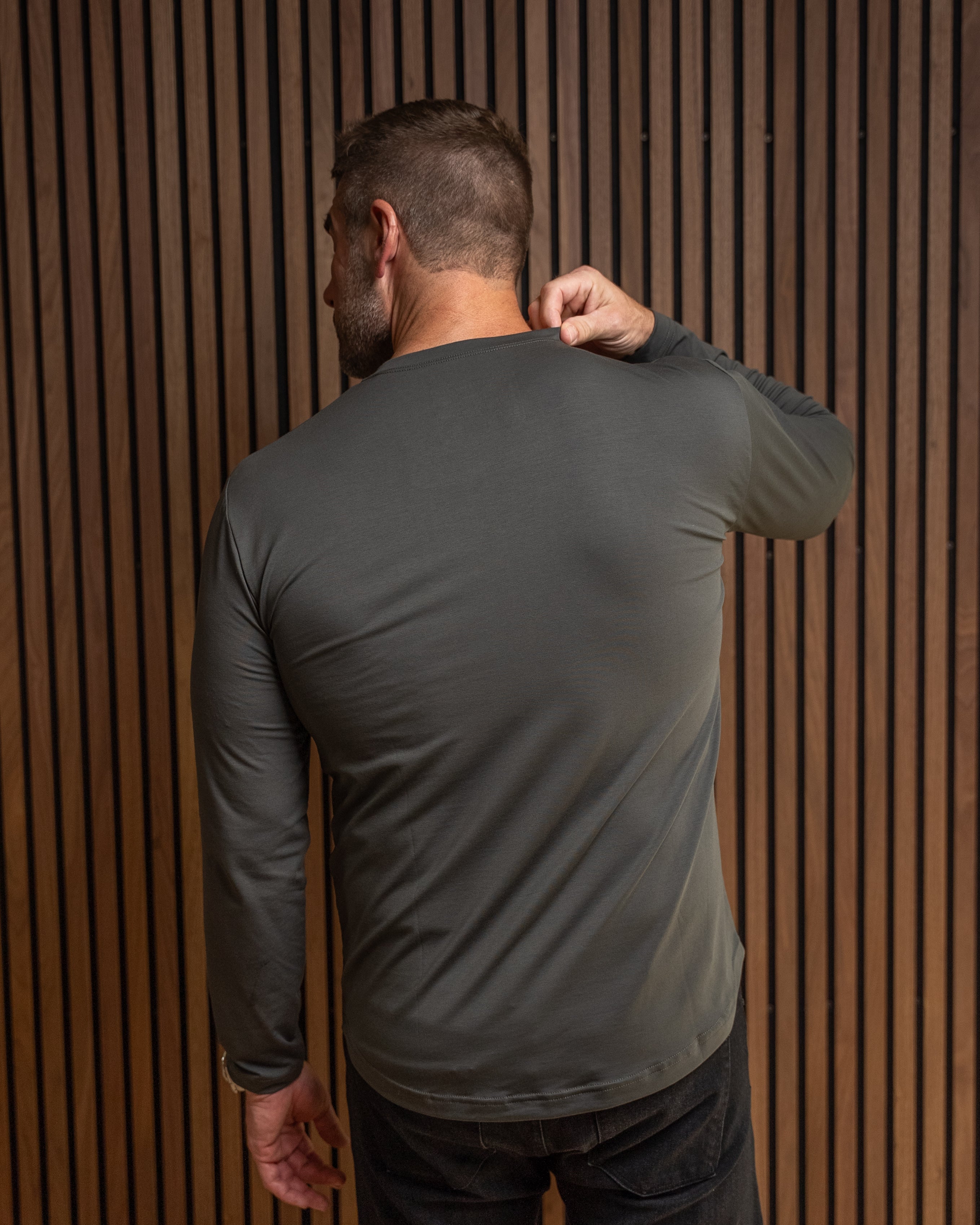 A man viewed from behind wearing a dark gray long-sleeve athletic shirt and dark jeans, standing against a brown vertical slat wall.