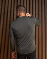 A man viewed from behind wearing a dark gray long-sleeve athletic shirt and dark jeans, standing against a brown vertical slat wall.