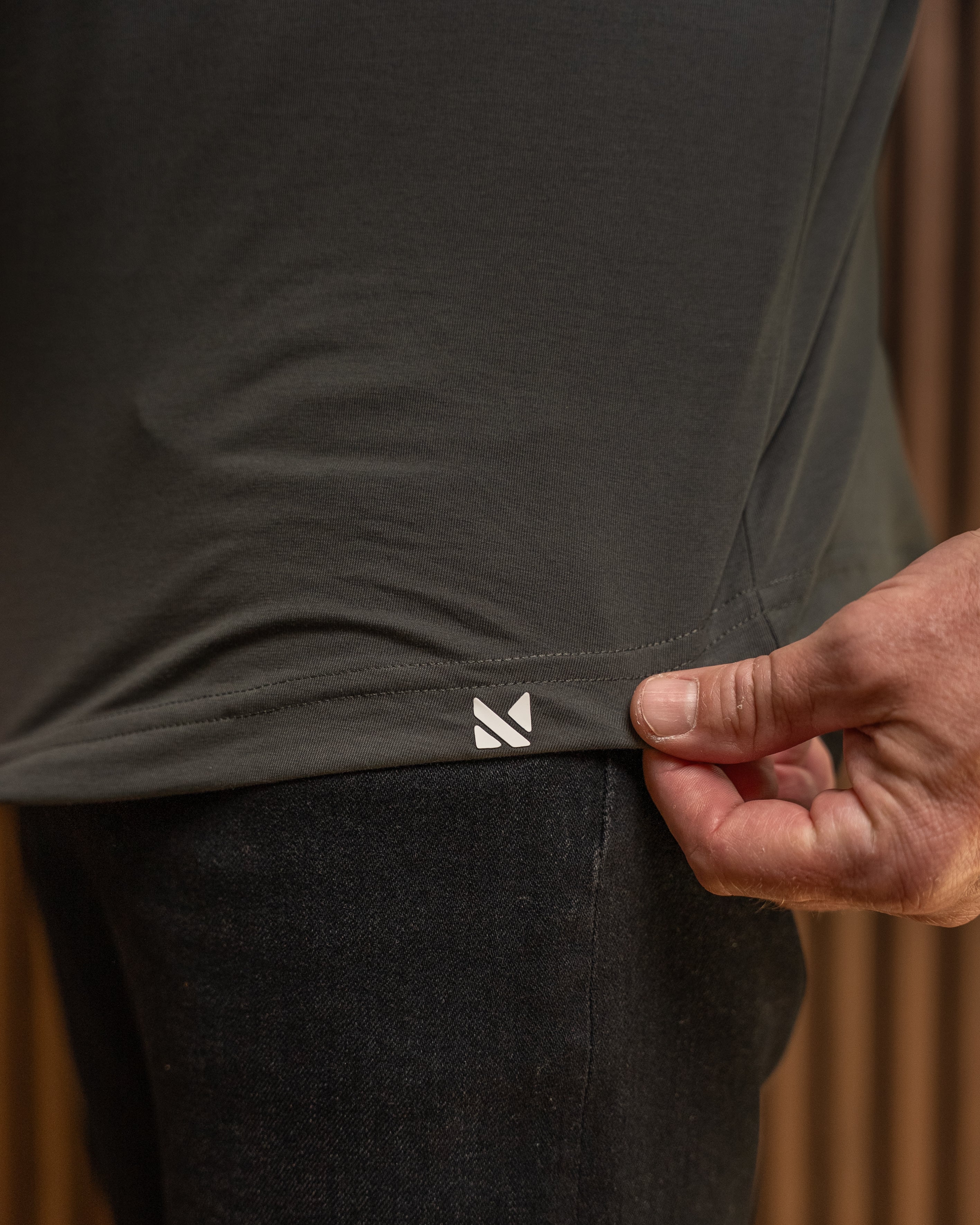 Close-up of a person wearing a dark charcoal gray t-shirt and black pants, pulling the hem of the t-shirt to reveal a small white geometric logo. The background is a warm, blurred brown wood panel.