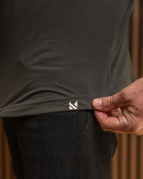 Close-up of a person wearing a dark charcoal gray t-shirt and black pants, pulling the hem of the t-shirt to reveal a small white geometric logo. The background is a warm, blurred brown wood panel.