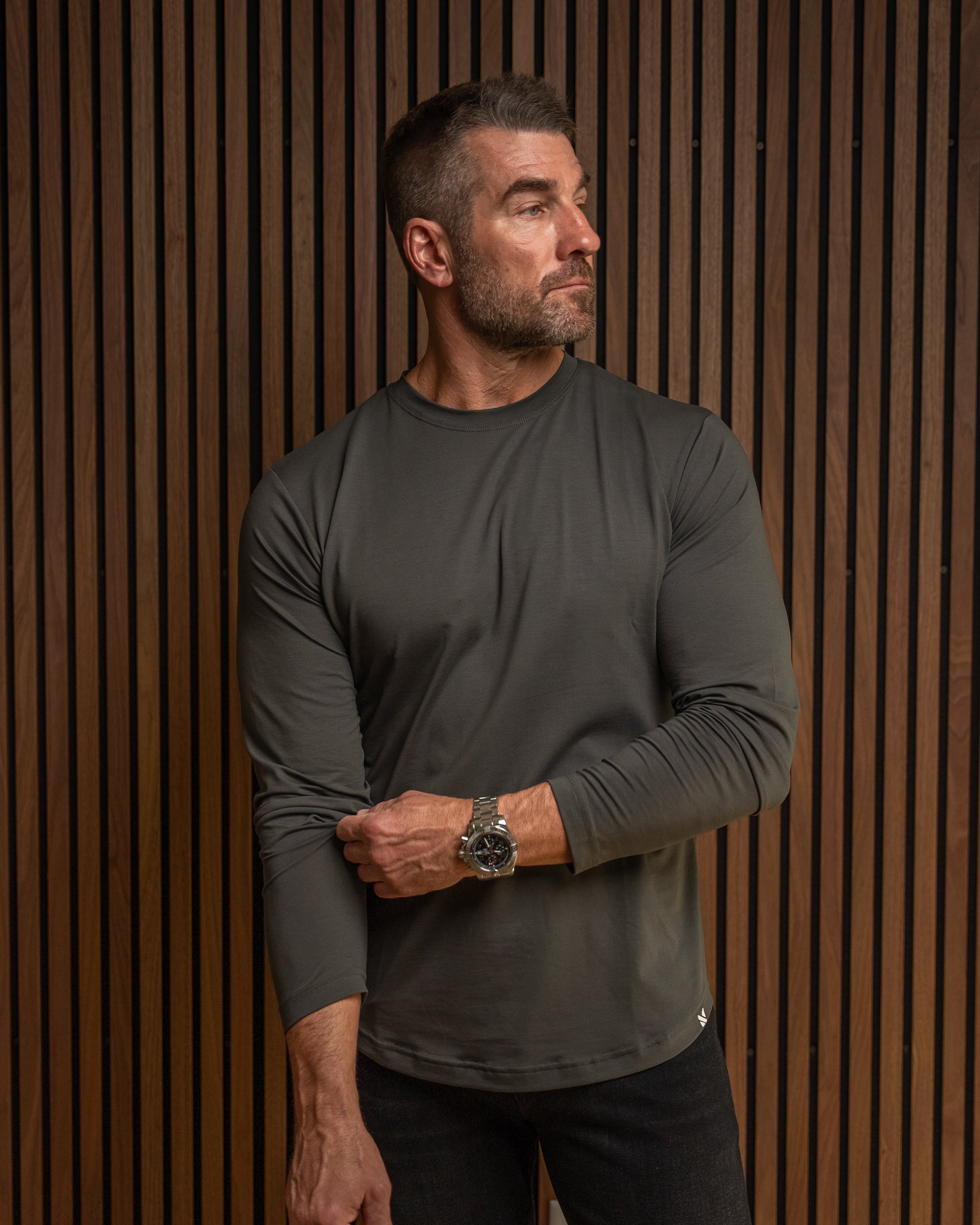A man wearing a dark gray long-sleeve athletic shirt and black pants, standing against a brown wood slat wall.