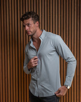 A man wearing a light gray long-sleeve button-down shirt and dark jeans, posing against a brown vertical slat wall.