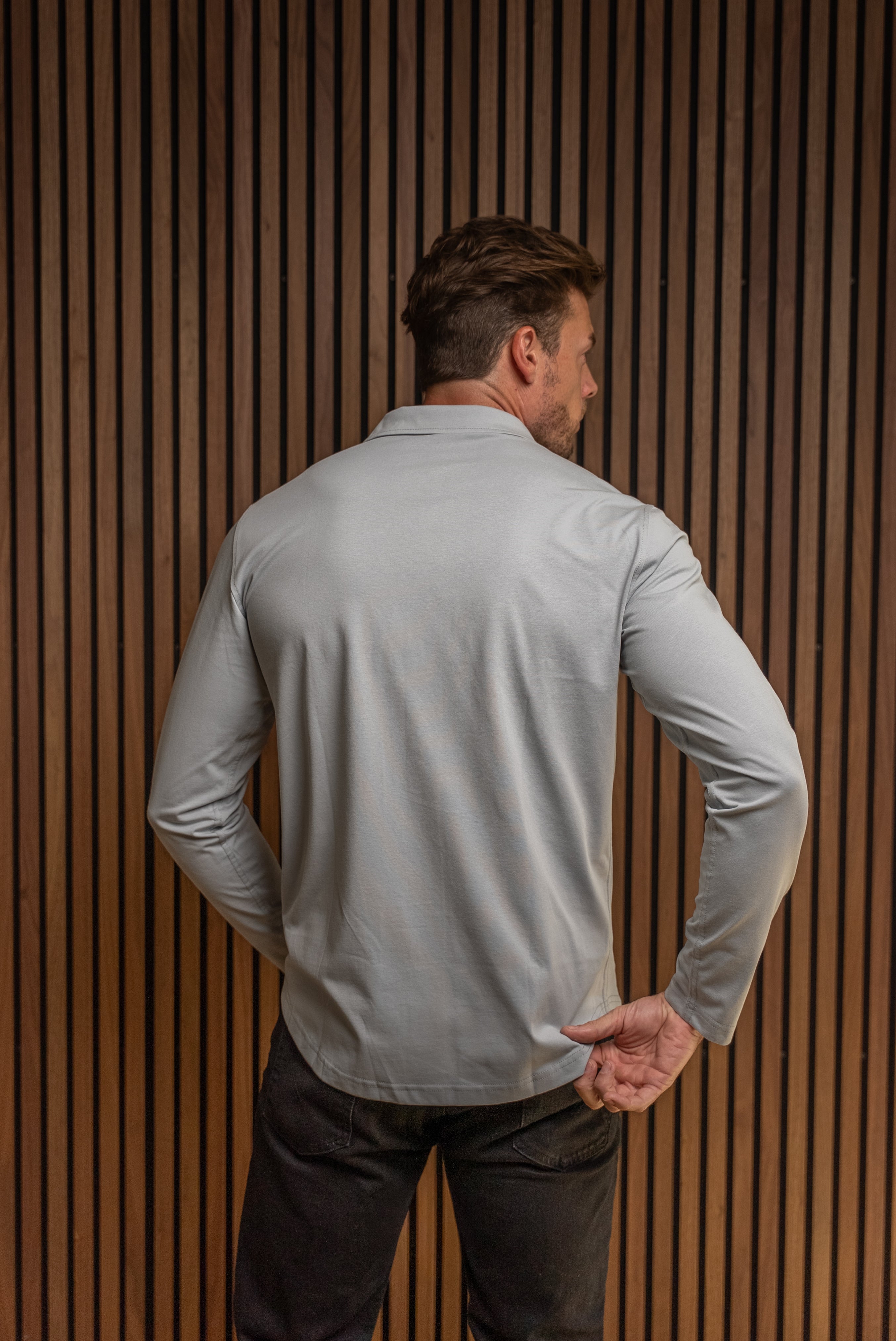 A man viewed from behind wearing a light blue long-sleeve performance shirt and dark pants, standing in front of a vertical wood slat wall.