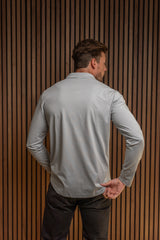 A man viewed from behind wearing a light blue long-sleeve performance shirt and dark pants, standing in front of a vertical wood slat wall.