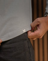 Close-up of a person wearing a light blue knit top and dark charcoal pants, pulling the hem of the top slightly to reveal a small white geometric logo.