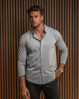 A man wearing a very light blue/grey button down against a walnut wood background with black pants and a silver watch.