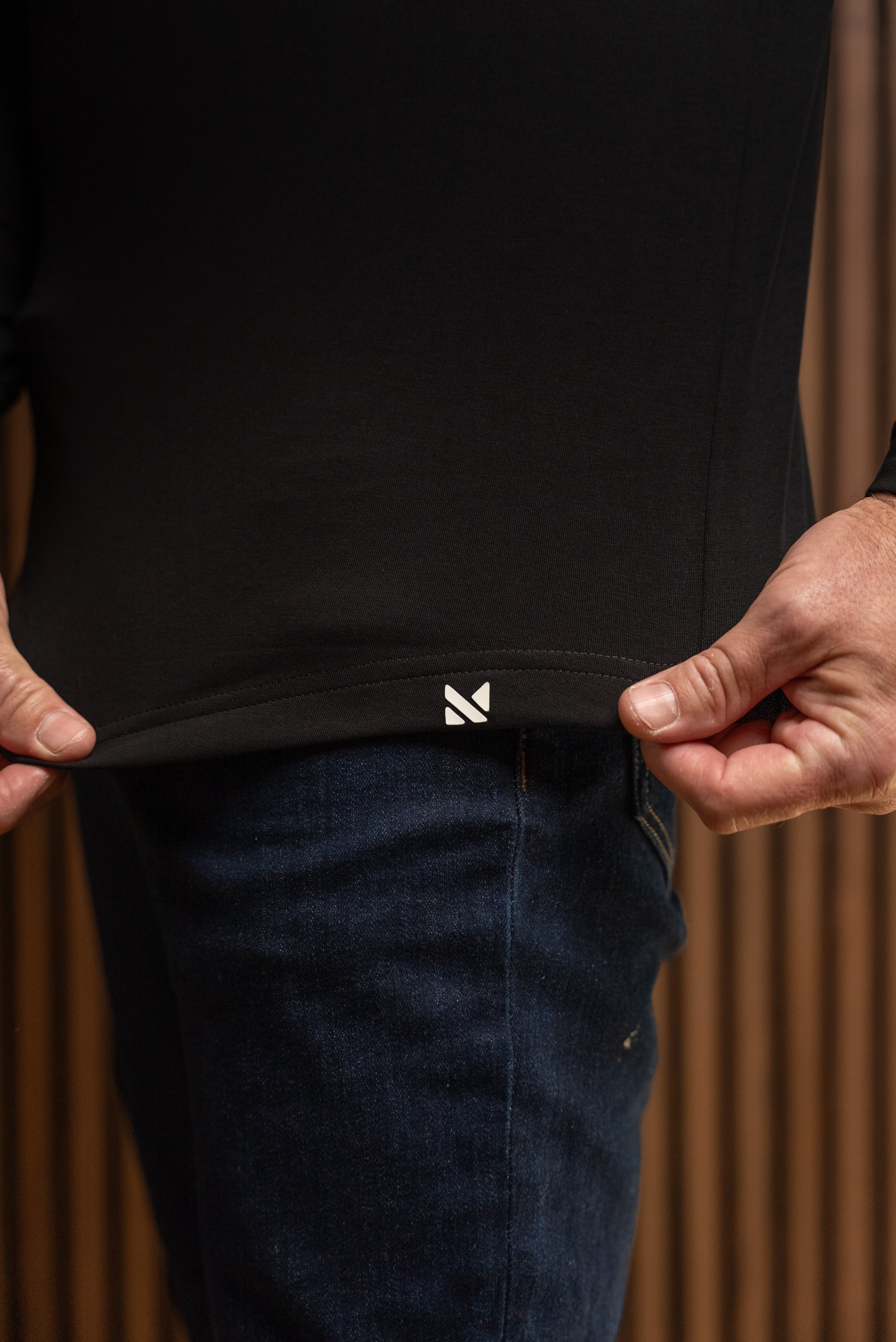 Close-up of a person wearing a black long-sleeve top and dark blue jeans, pulling the hem of the shirt slightly outward with both hands. A small white geometric logo is visible near the bottom hem.