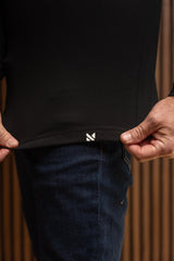 Close-up of a person wearing a black long-sleeve top and dark blue jeans, pulling the hem of the shirt slightly outward with both hands. A small white geometric logo is visible near the bottom hem.
