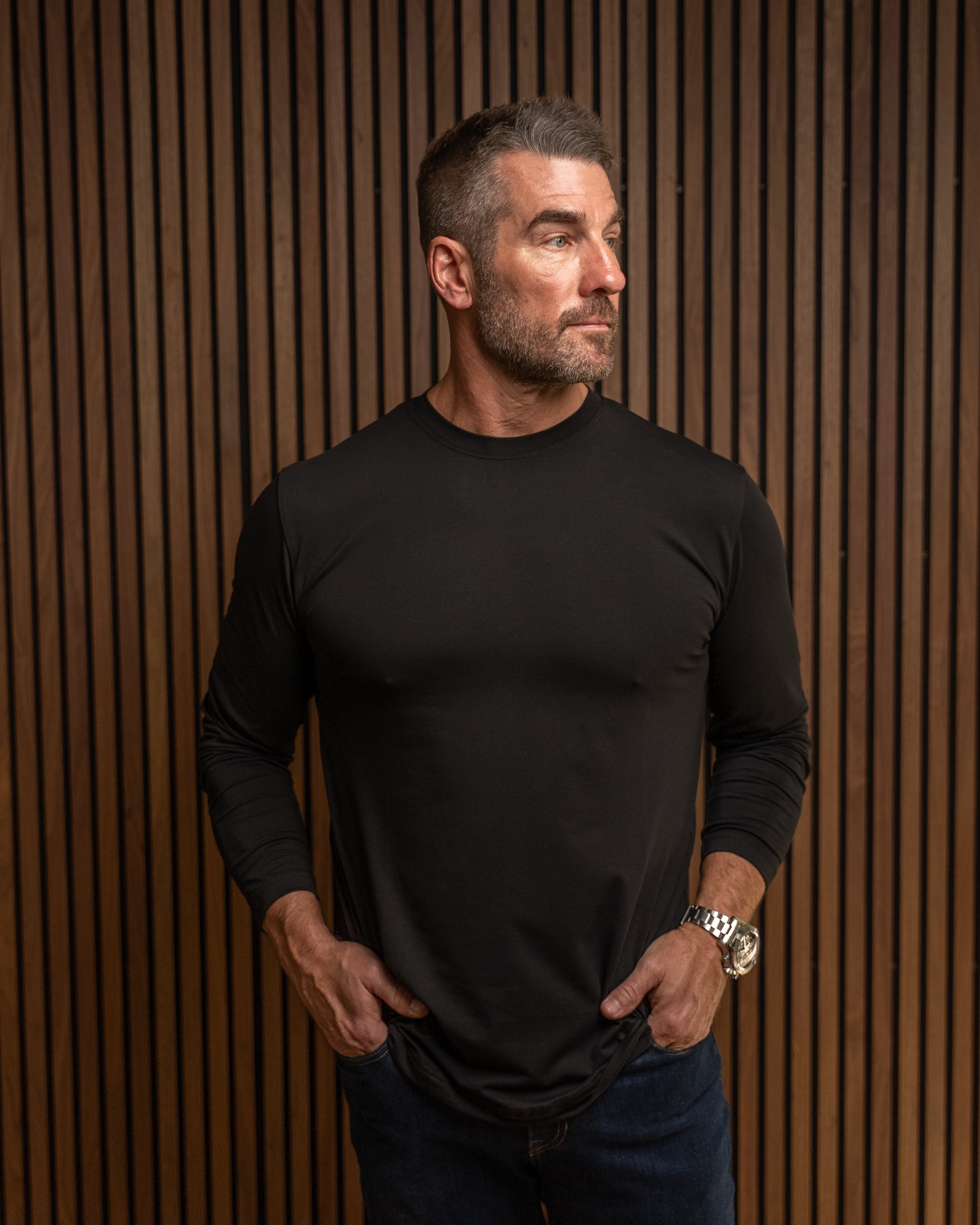 A man wearing a black long-sleeve crewneck shirt and dark blue jeans, standing against a brown vertical slat wall.