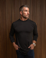 A man wearing a black long-sleeve crewneck shirt and dark blue jeans, standing against a brown vertical slat wall.
