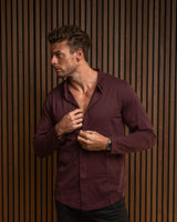 A man wearing a long-sleeve burgundy button-down shirt and black pants, standing against a brown vertical slat wall and buttoning his shirt.