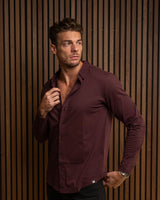 A man wearing a long-sleeve burgundy button-down shirt and black pants, posing against a vertical brown wood slat wall.