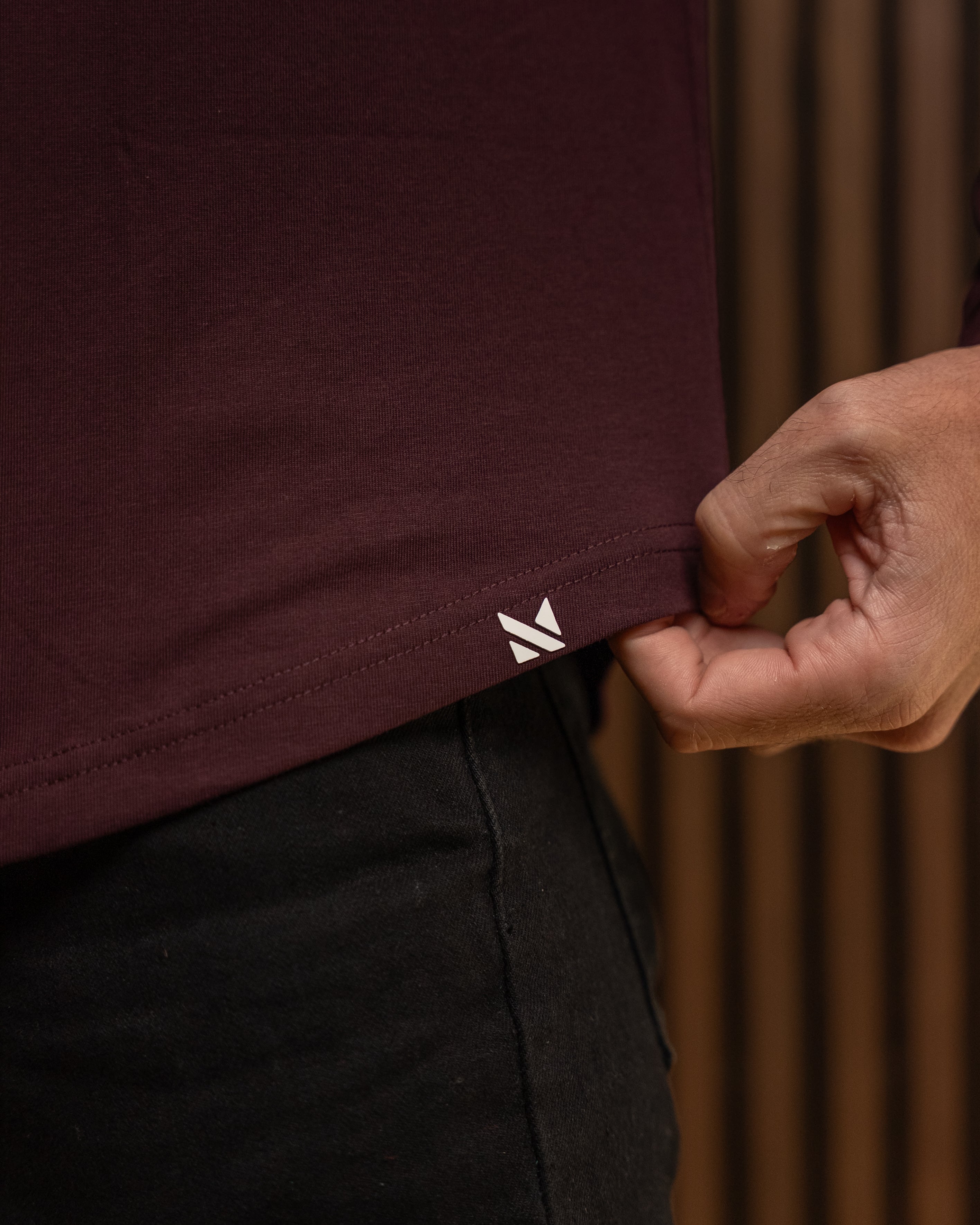 Close-up of a person wearing a dark burgundy t-shirt and black pants, pulling the hem of the shirt to reveal a small white embroidered logo. The background is a warm, brown textured wall.
