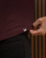 Close-up of a person wearing a dark burgundy t-shirt and black pants, pulling the hem of the shirt to reveal a small white embroidered logo. The background is a warm, brown textured wall.