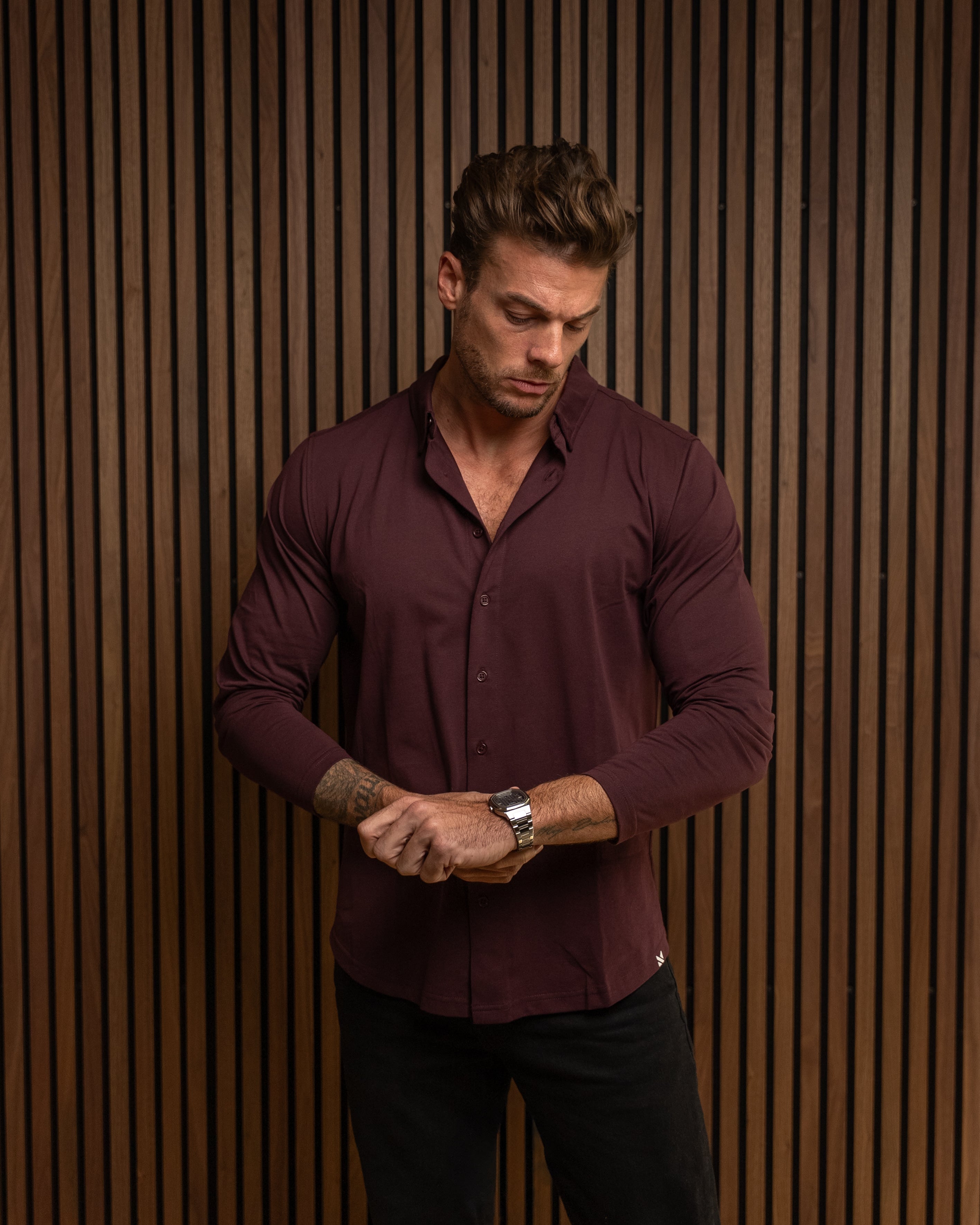 A man wearing a long-sleeve burgundy button-down shirt and black pants, standing in front of a brown vertical slat wall and looking down at his wrist.