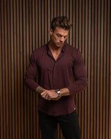 A man wearing a long-sleeve burgundy button-down shirt and black pants, standing in front of a brown vertical slat wall and looking down at his wrist.