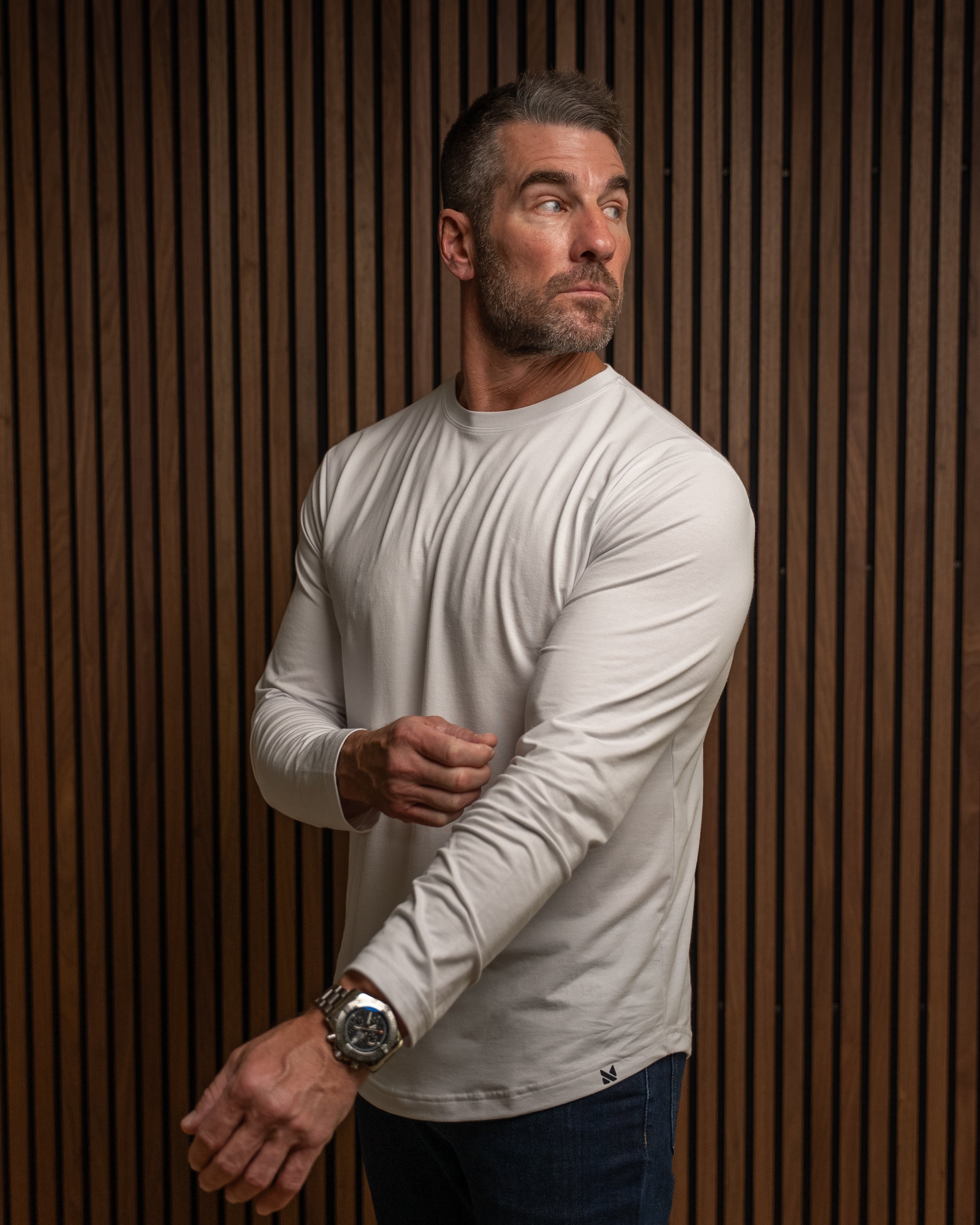 A man wearing a light beige long-sleeve crewneck shirt and dark blue jeans, standing against a brown wood slat wall.