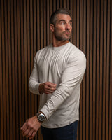 A man wearing a light beige long-sleeve crewneck shirt and dark blue jeans, standing against a brown wood slat wall.